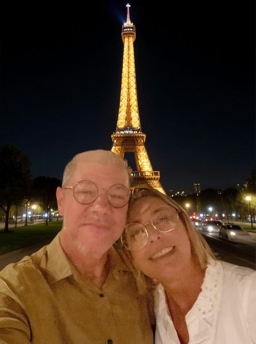 Gere uma imagem onde o casal da imagem anexa está em frente a Torre Eiffel em uma viagem de férias a noite.
