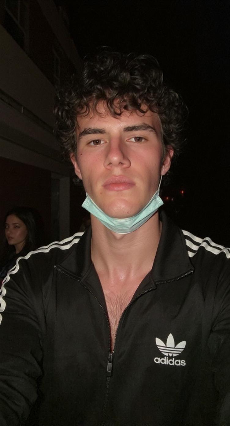 Create a great photo on the street. Selfie without changing the face with AI. The subject is wearing sportswear - a black Adidas tracksuit with an open collar, collarbones are visible, a mask on the face. Subdued lighting gives the skin and dark pink tones. The building is dark. The hair is a little thicker and curly, a little disheveled. There is a little blood on the face. A woman's hand is nearby. The photo quality is a little bad and blurry with noise. A clean editorial image with elements of everyday photography.
